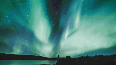 The aurora borealis displays streaks of green and blue light across a starry night sky above a silhouetted landscape.