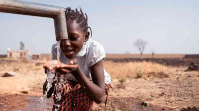 A young girl collects water from a metal tap outdoors in a dry, rural area, holding her hands under the flowing water.