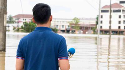 A person in a blue shirt holding a microphone stands facing a flooded urban area with buildings partially submerged in water.