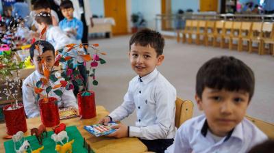 Children sit at a table in a classroom, surrounded by crafts and colorful decorations. Three boys in white shirts focus on activities.