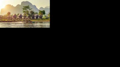 A river with two people in a small boat passes by stilted houses and palm trees, with misty mountains in the background.