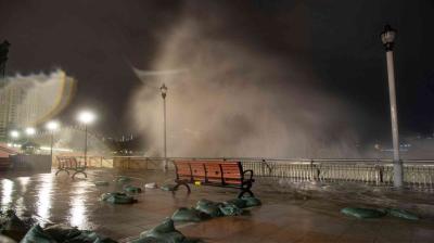 Large waves crash over a waterfront promenade at night, soaking benches and sandbags, with city lights and street lamps visible in the background.