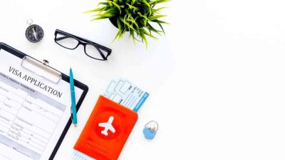 A visa application form on a clipboard is shown with a pen, eyeglasses, a compass, a plant, a passport holder, boarding passes, and a pin on a white surface.