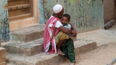 Un homme âgé, coiffé d'une casquette blanche et d'un tissu à motifs, est assis sur des marches extérieures, tenant dans ses bras un jeune enfant qui se couvre le visage d'une main. Le décor paraît délabré et rustique.