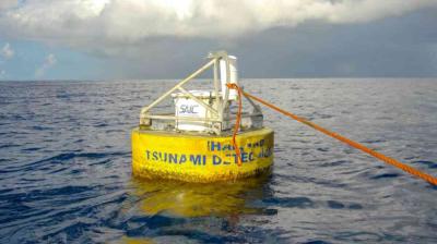 A yellow tsunami detection buoy floats on the ocean, anchored by an orange rope, with cloudy sky in the background.