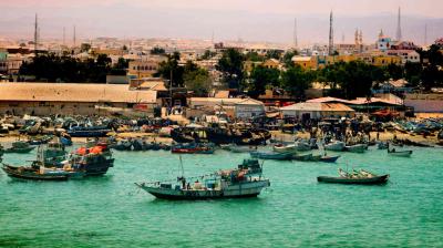 Fishing boats are anchored near the shore with a busy coastal town and distant mountains in the background under a hazy sky.