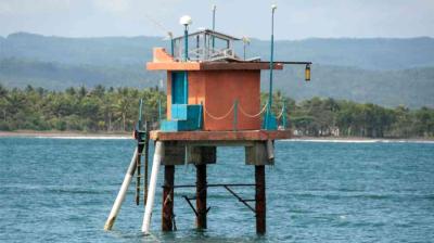 An orange and blue stilted structure stands above the water near a coastline, with a ladder, railings, and equipment on the roof.