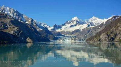 Snow-covered mountains and a glacier reflected in a calm, clear lake under a blue sky.