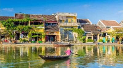 A person wearing a conical hat rows a small wooden boat on a calm river, with colorful old buildings and greenery lining the riverside under a clear blue sky.