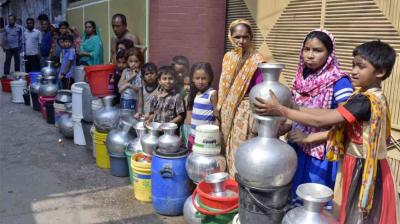 A group of people, including women and children, stand in line with metal and plastic containers, waiting to collect water on a street.