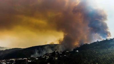 Smoke billows from a wildfire on a forested hillside near a residential area under a cloudy, orange-tinted sky.
