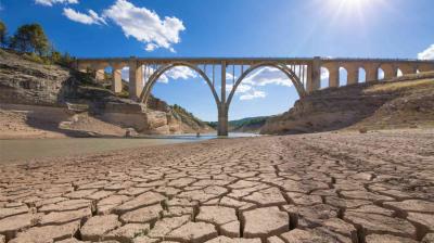 A stone arch bridge spans a river with visibly low water levels, surrounded by cracked, dry earth under a bright blue sky.