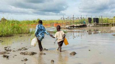 Two children walk through a muddy, waterlogged area carrying plastic containers, with green fields and makeshift structures in the background.