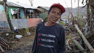 A man wearing an "I ❤️ Tacloban" shirt stands in front of a damaged home surrounded by debris.