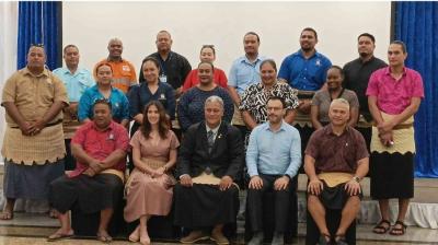 A group of twenty people, including men and women, pose in two rows. Some wear traditional Polynesian attire with woven mats. A plain backdrop is behind them.
