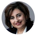 A person with dark, shoulder-length hair smiles while wearing pearl earrings and a dark blazer in a circular framed photo.