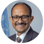 A man with glasses and a mustache, smiling, wearing a suit and tie, with a United Nations flag in the background.