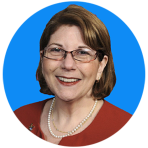 A woman with glasses, short brown hair, and a red blazer smiles against a bright blue background. She is wearing a pearl necklace.