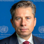 A man in a suit and tie stands in front of a blue background with United Nations logos and text.