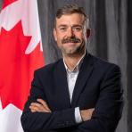 A man in a navy blazer stands with arms crossed in front of a Canadian flag and dark curtains.