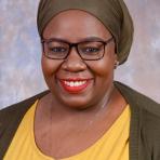A woman wearing glasses, a green headscarf, and a yellow top smiles at the camera against a neutral, mottled background.