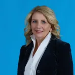 A woman with blonde hair wearing a dark blazer and white blouse stands against a solid blue background, looking at the camera.