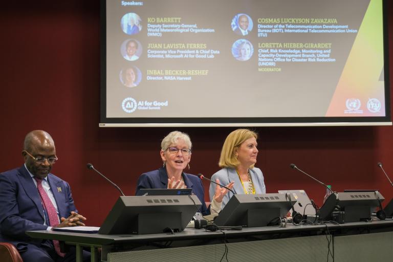 Three individuals are seated on a panel at a conference. A large screen behind them displays their names and titles, with "AI for Good" and other logos visible.