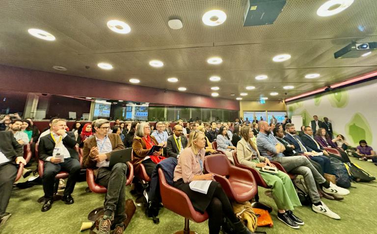 A seated audience attentively listening to a speaker in a conference room with a burgundy and beige color scheme. The room is equipped with overhead lighting and a projector.