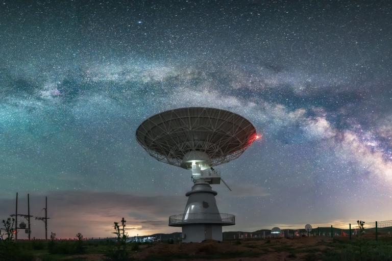 A large radio telescope dish is silhouetted against a star-filled sky and the Milky Way galaxy.